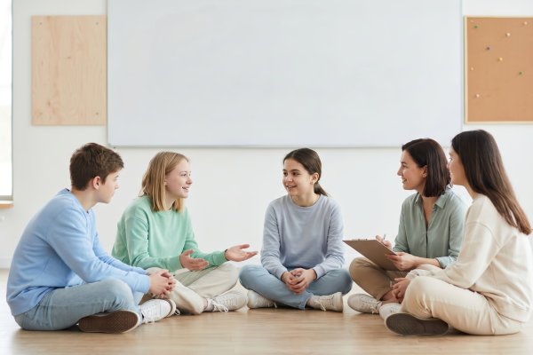 Teenagers and adults sitting in a circle on the floor of a bright classroom, in front of a whiteboard